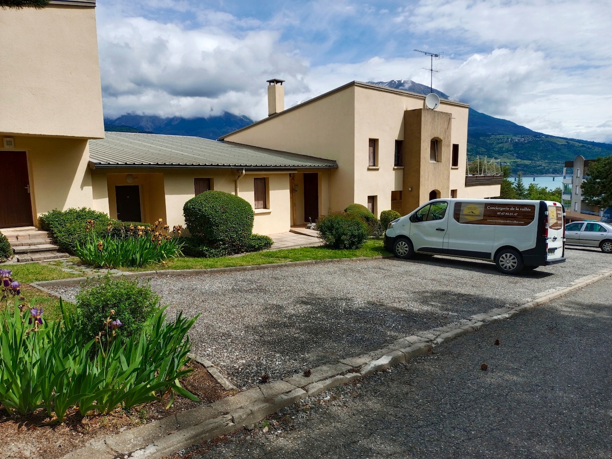 The exterior of a two-story building is shown, surrounded by low-maintenance greenery and a gravel path. A vehicle is parked nearby, and distant mountains can be seen under a partly cloudy sky. A lake is visible in the background, suggesting proximity to water.