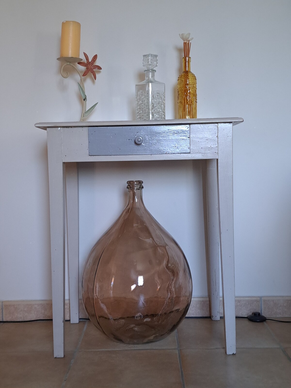 A small, light-colored table features a single drawer and is complemented by decorative items. A large, rounded glass container rests on the floor below, and various glass bottles with differing shapes and colors are displayed above, alongside a candle holder and a flower decoration.