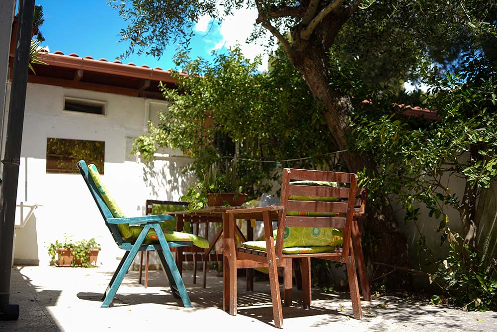An outdoor seating area features a wooden table surrounded by two chairs, one with a vibrant green cushion and the other painted in blue. Lush greenery adds a natural setting, complemented by a sunny sky and terracotta roof overhead.