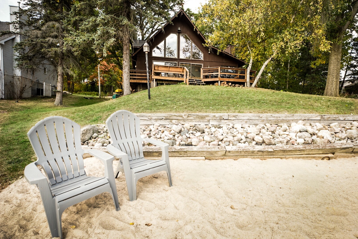 Two gray adirondack chairs are placed on a sandy area, facing a grassy slope leading to a spacious brown cabin. The cabin features a large deck with a set of stairs and is surrounded by trees, creating a serene outdoor setting.