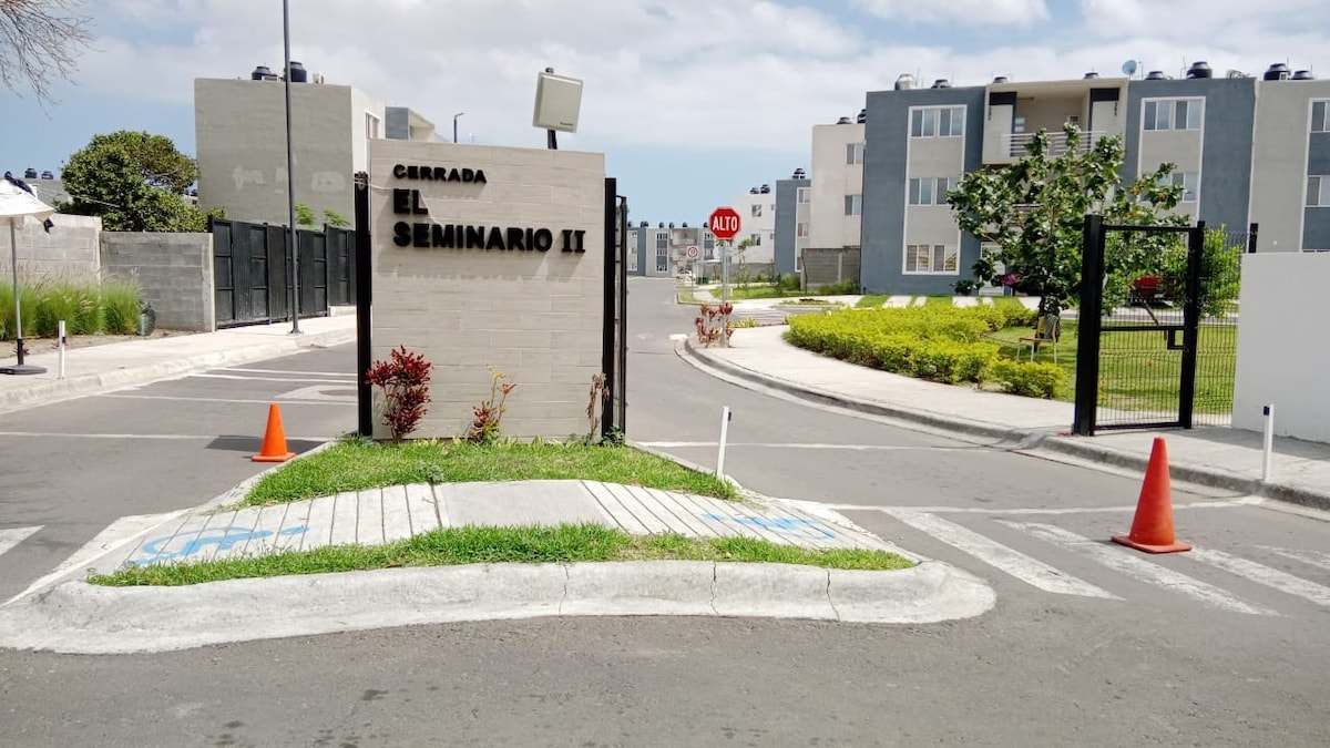 The entrance to the gated community is marked by a sign reading 'Cerrada El Seminario II'. Lush greenery lines the paths, and traffic cones are positioned near the entry, indicating careful management of the space. A clear blue sky offers a bright backdrop.
