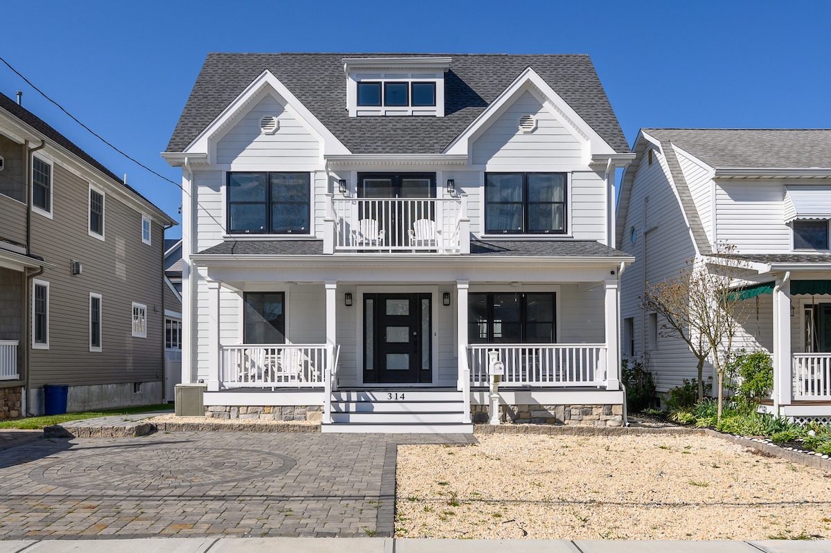 A modern two-story house is showcased, featuring a light-colored facade and large windows. A spacious front porch is adorned with chairs, while a paved walkway leads up to the entrance. The front yard is landscaped with gravel and minimal greenery, allowing for easy maintenance.