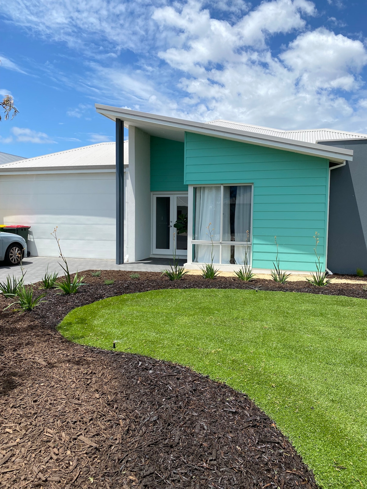 The exterior of the apartment features a modern design with a turquoise facade and large front windows. A well-maintained garden showcases a mix of vibrant green grass and mulch, along with low-growing plants arranged along the pathway leading to the entrance.
