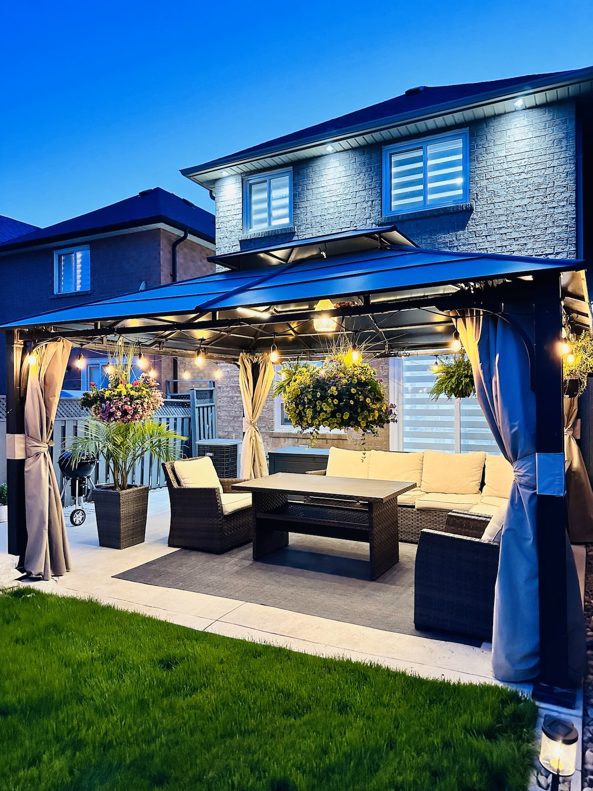 An inviting backyard patio showcases a cozy seating area under a gazebo, adorned with hanging plants and string lights. Softly illuminated in the evening, the space invites relaxation and outdoor gatherings with a low coffee table and comfortable furnishings surrounded by well-maintained green grass.