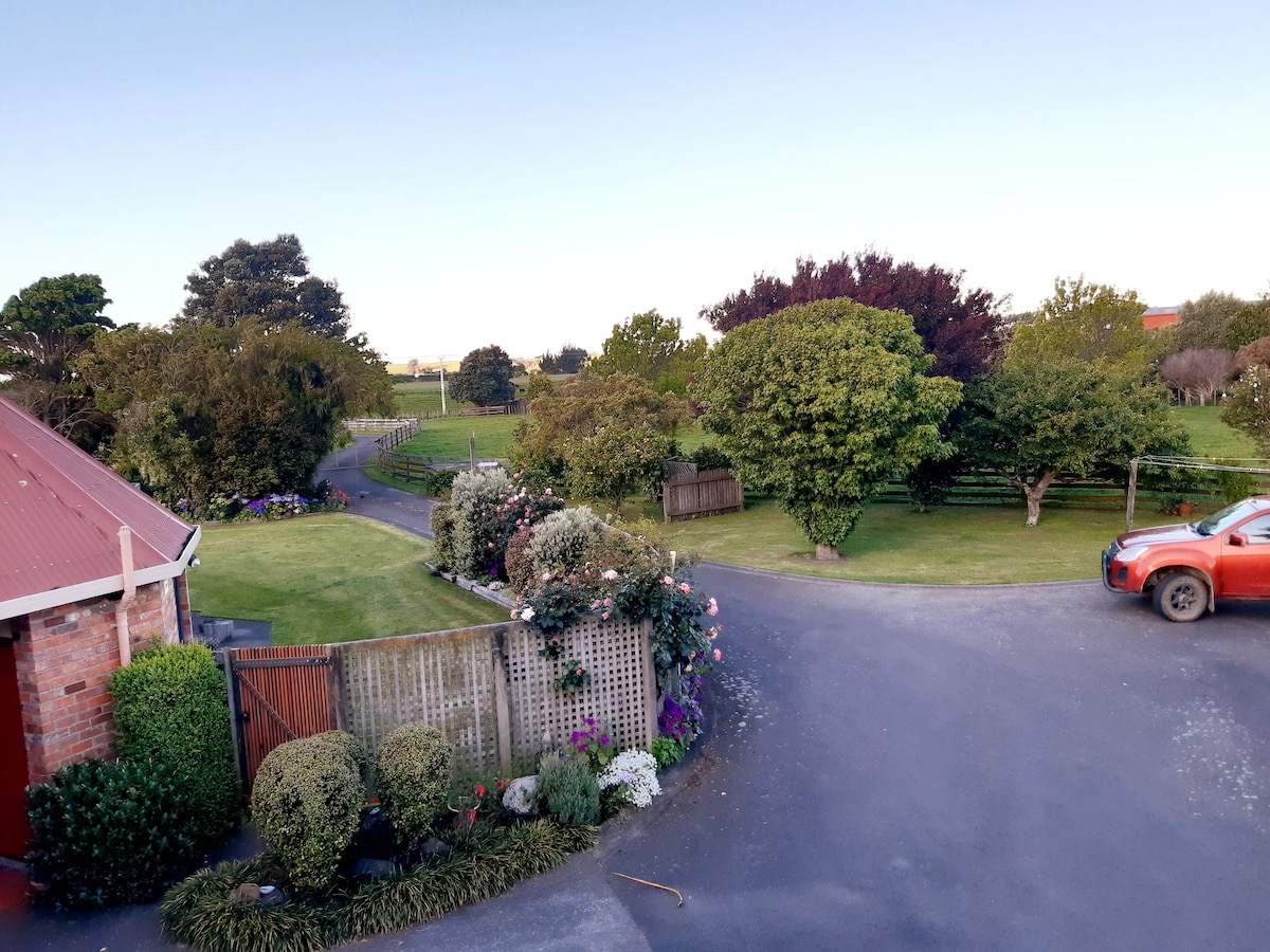 A scenic view from above captures the landscaped gardens surrounding the property, featuring a winding driveway and lush greenery. Trees and flowering plants add vibrant touches to the serene rural atmosphere, with a glimpse of nearby fields in the distance.