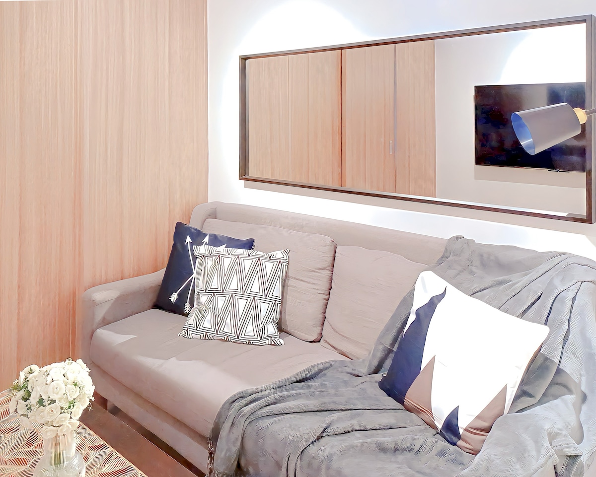 A comfortable sofa is presented, adorned with decorative pillows in varying patterns. A soft throw blanket drapes over one side. A mirror above the sofa reflects subtle light, while a wall-mounted television is positioned nearby, creating a cozy living space.