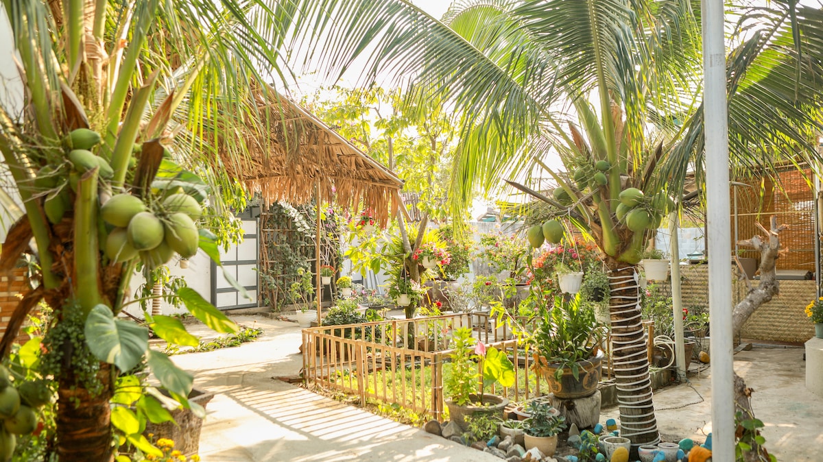 A sunlit outdoor garden area is framed by tall palm trees and lush greenery. A bamboo-roofed structure is visible in the background. A fenced garden featuring various plants, flowers, and decorative stones adds to the tranquil atmosphere of the space.