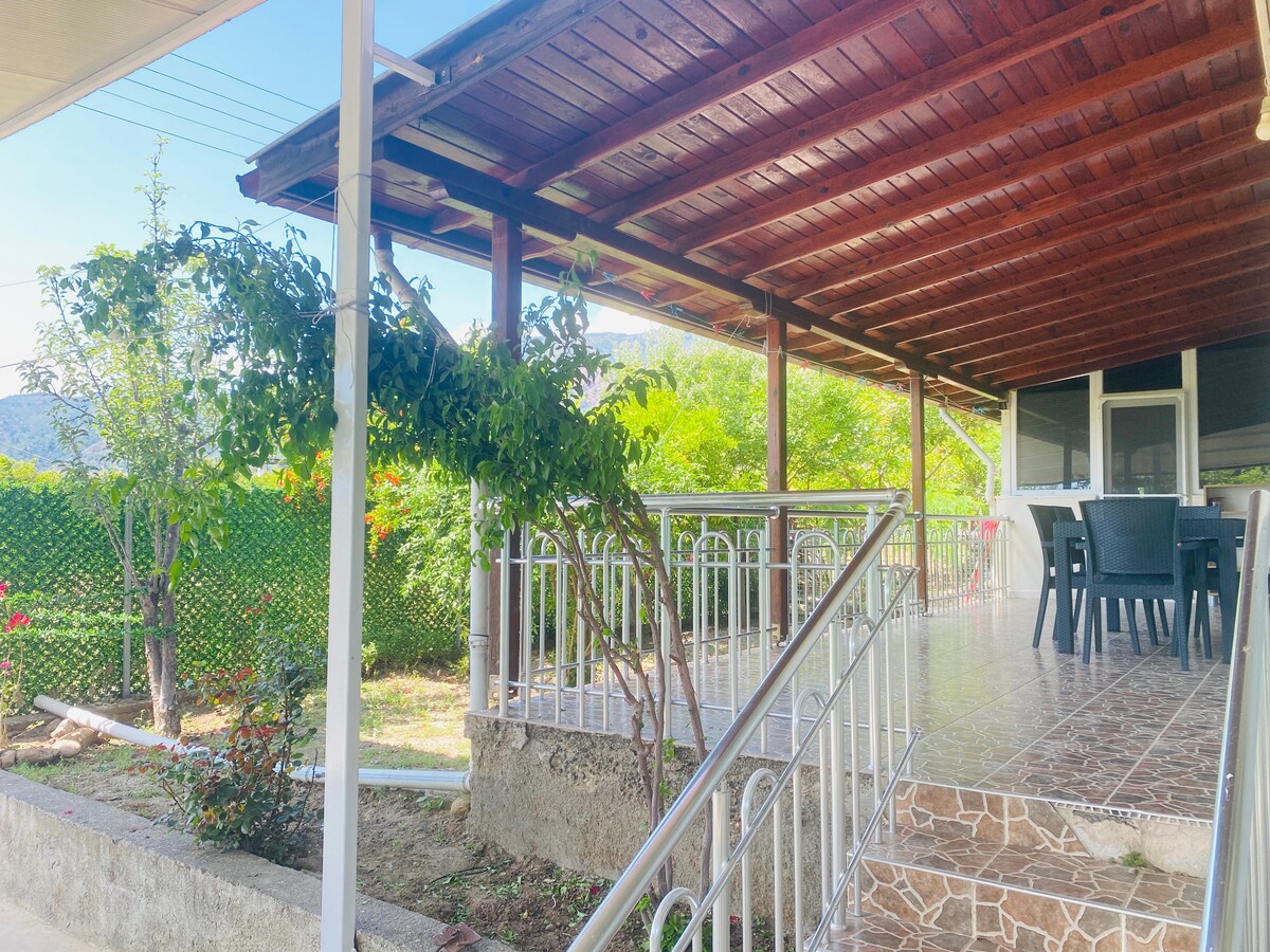 A covered patio area features a wooden ceiling supported by beams. A selection of outdoor furniture, including a table and chairs, is arranged for relaxation. Lush greenery surrounds the space, offering a peaceful view of the garden, while the tiled flooring adds a touch of elegance.