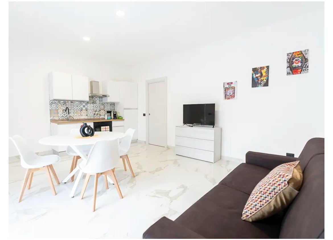 A bright living area features a dark sofa beside a white dining table with four chairs. The modern kitchen is visible in the background, with light cabinetry and patterned backsplash. A television is placed on a low dresser, complemented by two pieces of wall art.