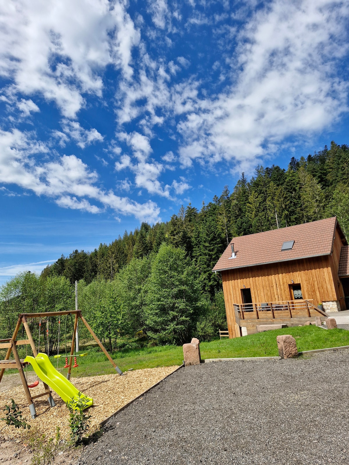 An outdoor area is depicted with a wooden house surrounded by lush greenery and towering trees. A playground is visible, featuring a yellow slide and swings. The sky is mostly clear with scattered clouds, enhancing the serene, natural setting.
