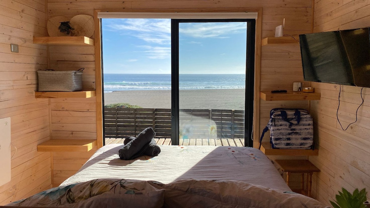 A bright interior is visible, featuring a spacious bed with soft linens. Large windows frame a view of the beach and ocean, allowing natural light to fill the room. Wooden shelves line the walls, offering storage and personal items. A wall-mounted TV is also present.