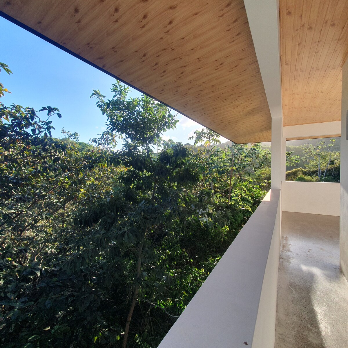 An upper terrace offers a view surrounded by lush greenery, with trees visible in the foreground. The ceiling features wooden panels, adding a natural touch to the space. The terrace is enclosed by a white railing, providing a sense of openness and tranquility.