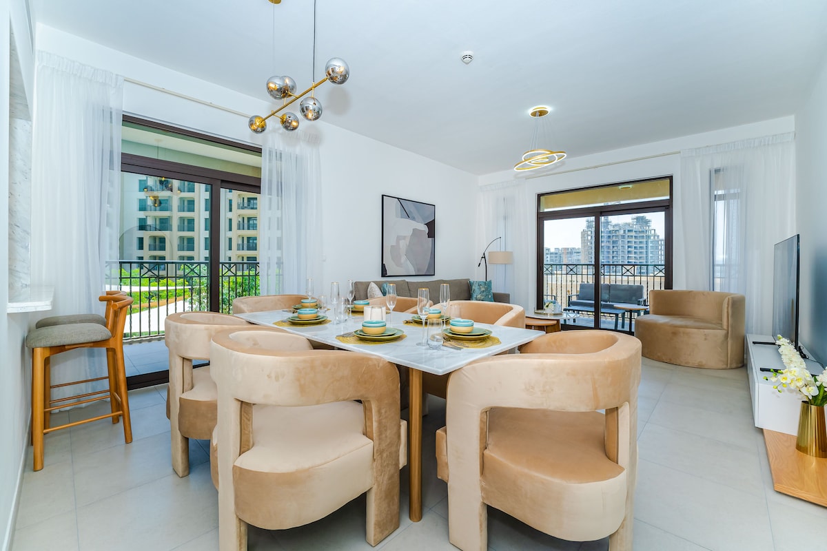 A dining area features a round marble table surrounded by plush beige chairs. Large windows allow natural light to fill the space, while a balcony view displays the community outside. An elegant light fixture hangs above, enhancing the modern design.