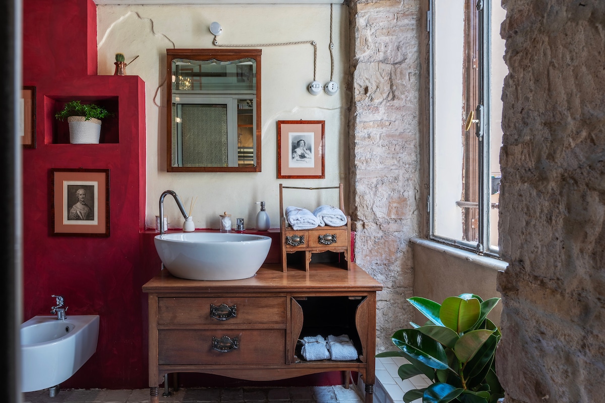 A stylish bathroom features a round sink mounted on a wooden vanity with a drawer and shelves for storage. A mirrored cabinet hangs above, and framed art pieces adorn the walls. Natural light streams in through a large window, complemented by green plants near the window.