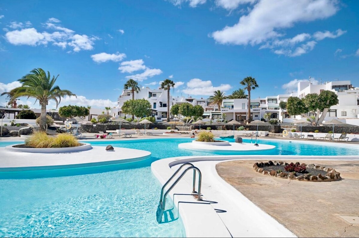 A large swimming pool is framed by white walkways and lush greenery, featuring gentle curves and varying depths. Palm trees provide shade, and lounge chairs are arranged around the pool area. Bright blue skies with scattered clouds create a vibrant atmosphere.