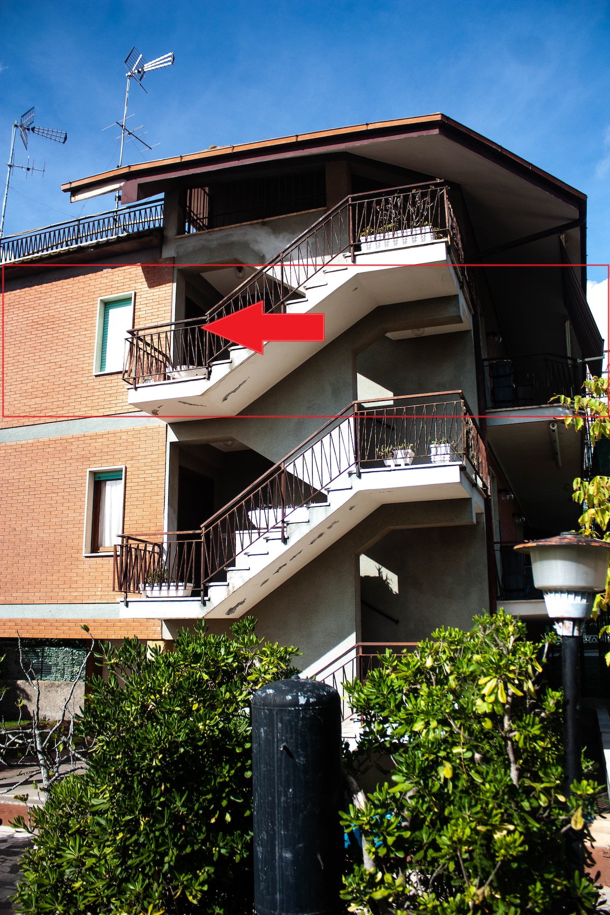 An exterior view of the building showcases a set of stairs leading to the second floor. The stairwell is flanked by a well-maintained garden area, with greenery visible in the foreground. The building features a combination of brick and stucco finishes.