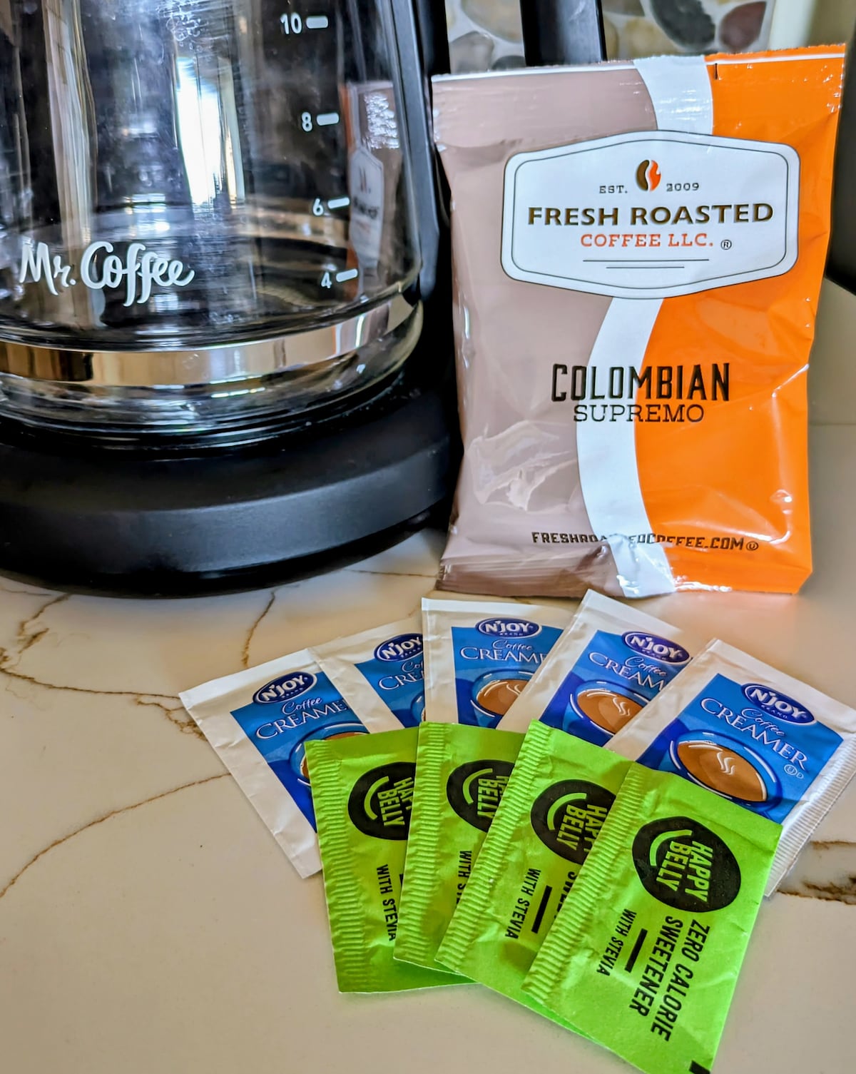 A coffee-making setup is displayed on a countertop, featuring a Mr. Coffee brewer and a bag of Colombian Supremo coffee. Several single-serve creamers and sweeteners are arranged in front, offering convenient options for guests.