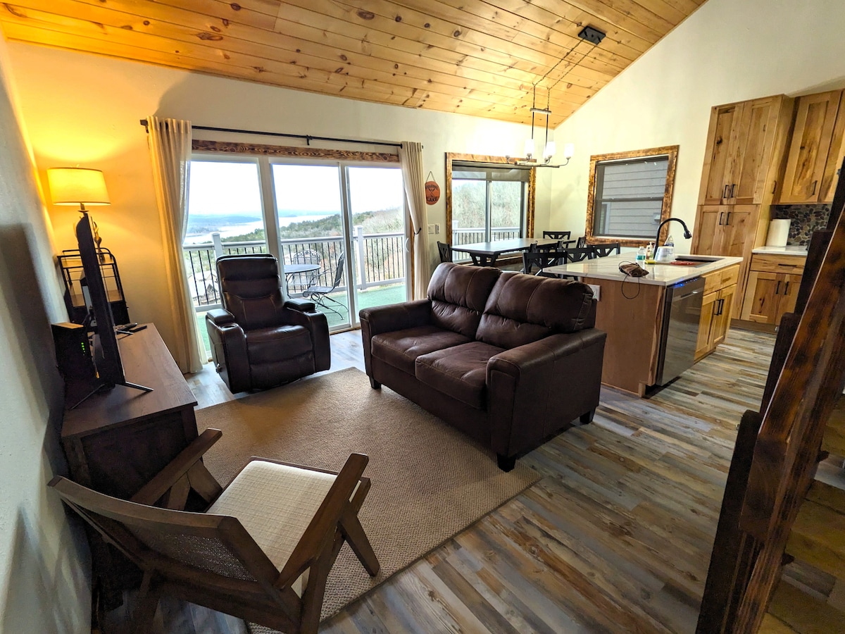 The living area features a comfortable brown leather sofa and a reclining chair facing a television. Large sliding doors allow natural light and offer views of the outdoor deck and surrounding landscape. A dining area with high-backed chairs is adjacent to the kitchen.