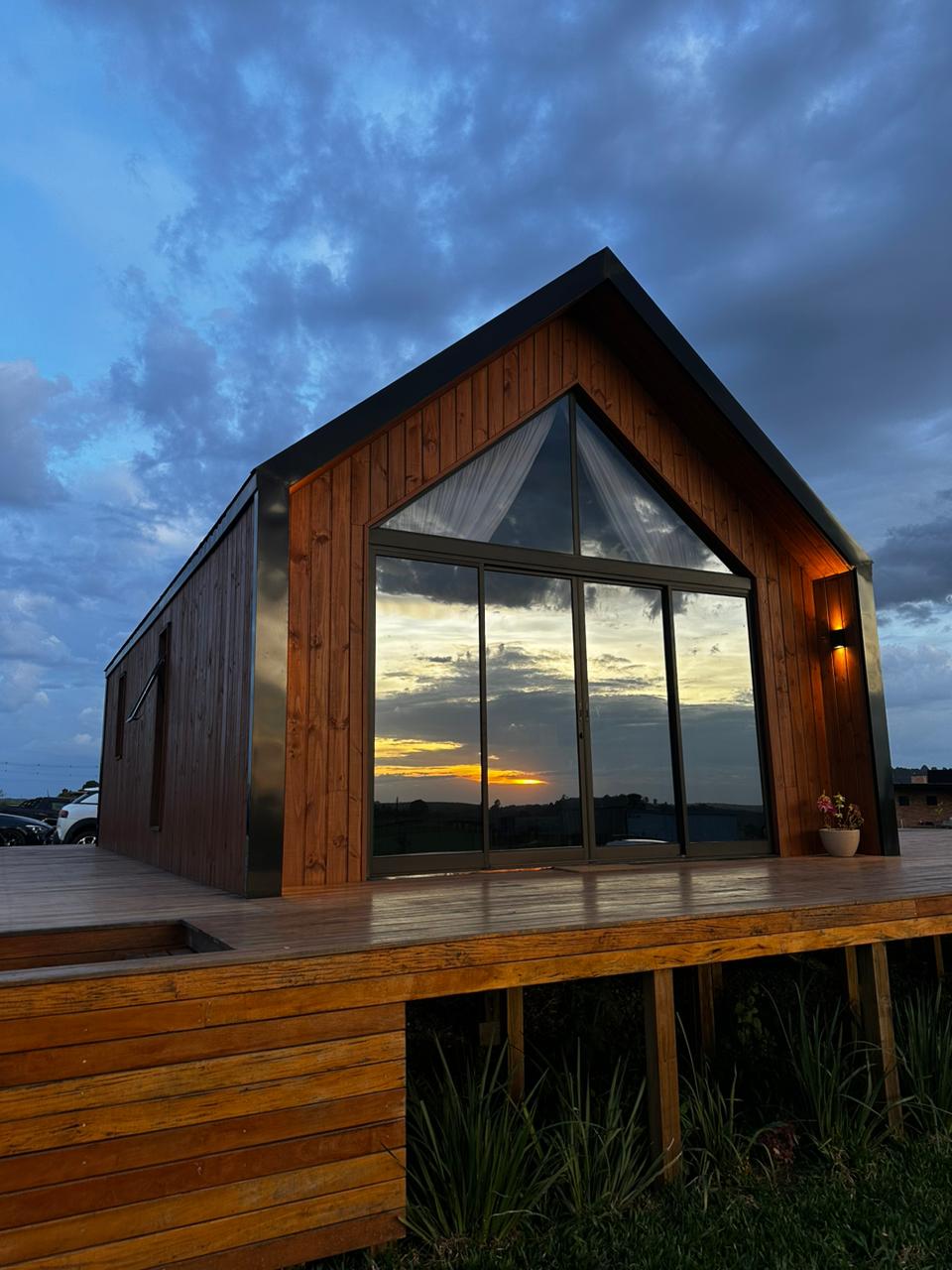 A modern cabin is showcased, featuring a striking triangular roof and large glass doors reflecting a colorful sunset. The wooden deck extends outward, surrounded by greenery, creating a harmonious connection with the natural environment.