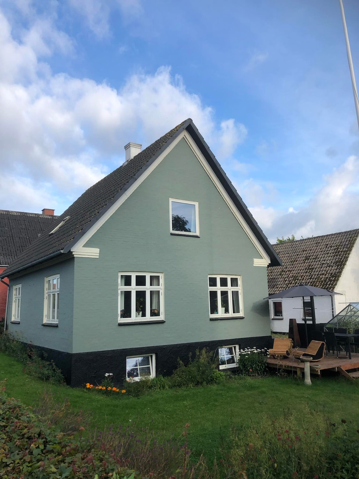 A charming two-story house is displayed with a sloped roof and inviting light blue exterior. Large windows allow natural light to fill the space. The surrounding garden features greenery and flowers, enhancing the outdoor appeal. Nearby structures complement the serene village atmosphere.
