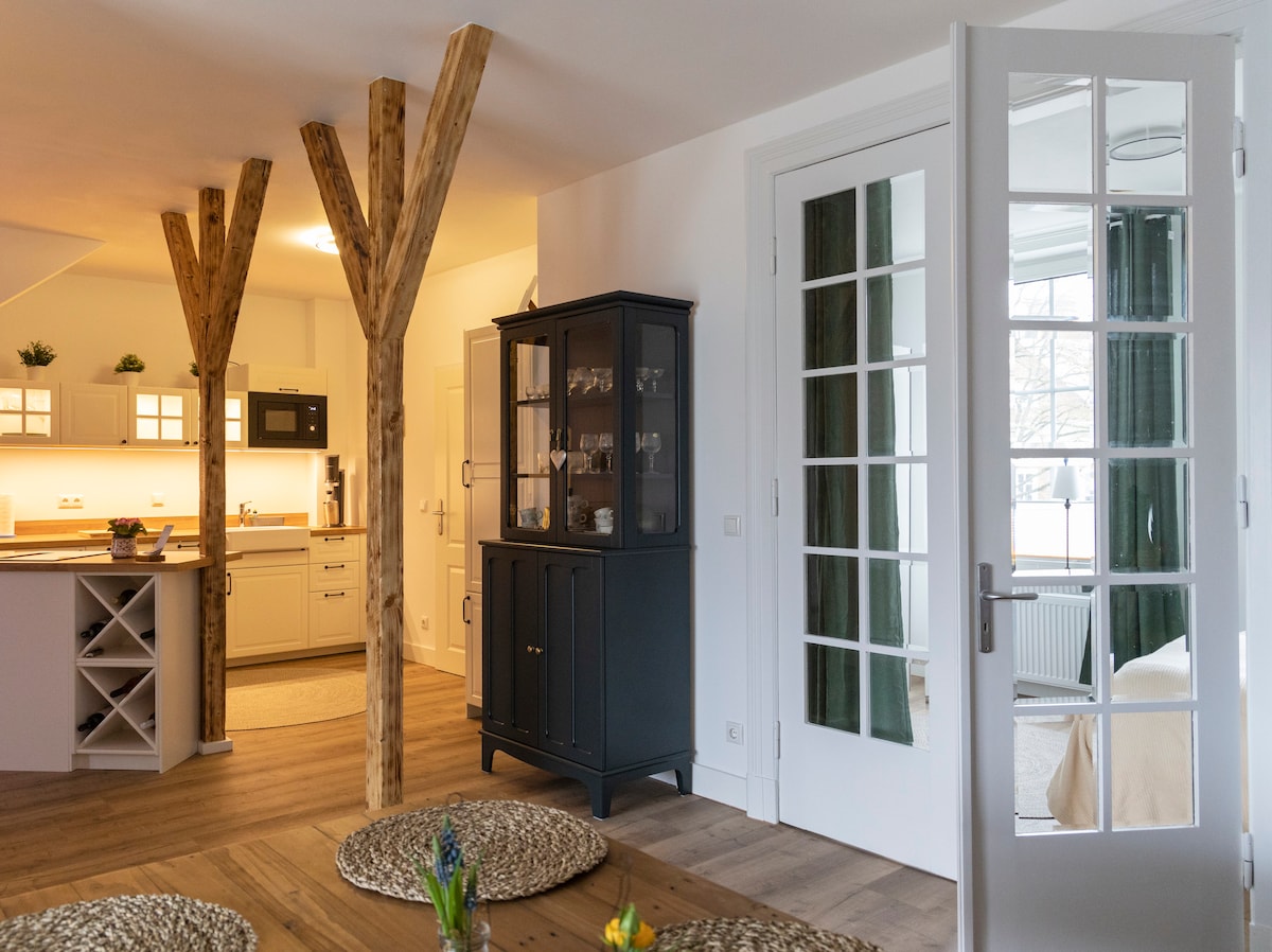 The image features a spacious living area with wooden beams accentuating the room. A black cabinet showcases glassware, while a door leads to another room. Natural light illuminates the space, highlighting the open kitchen area equipped with modern appliances.