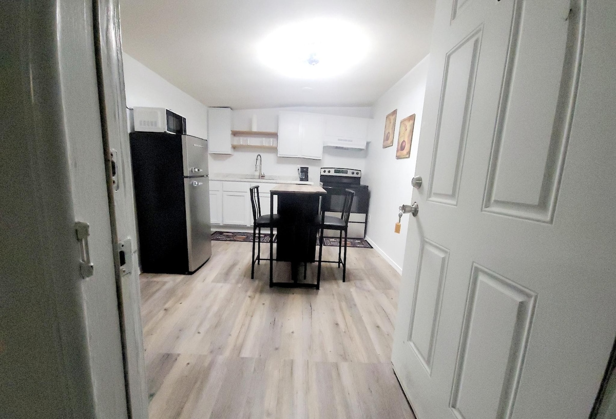 A well-lit kitchen area is visible, featuring modern appliances including a black refrigerator and white cabinetry. A dining table surrounded by four chairs is positioned centrally, with a stove and microwave in the cabinetry. Light wood flooring enhances the spacious feel.