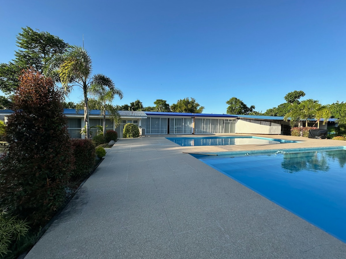 A spacious pool area is highlighted by clear blue waters, surrounded by well-kept landscaping. Palm trees and lush greenery enhance the serene environment, while a smooth stone pathway leads to the nearby building with large glass doors reflecting the sky.