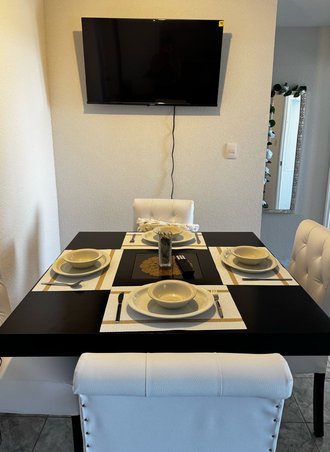 A dining area features a black table set for four, with white plates, utensils, and a central decorative piece. A television is mounted on the wall, while a mirror reflects the space. White upholstered chairs surround the table, offering a modern aesthetic.