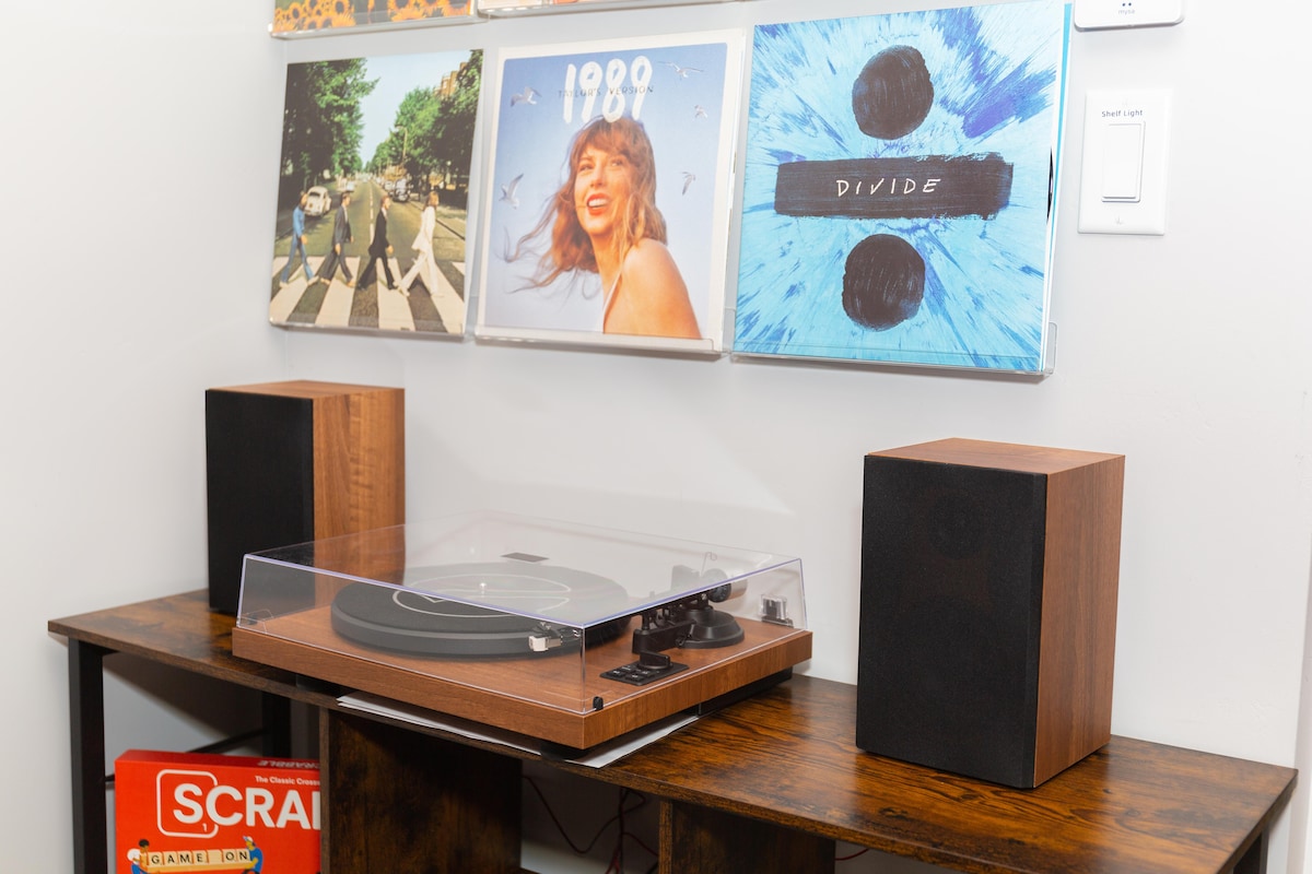 A stylish record player is placed on a wooden table, accompanied by two speakers on either side. Above the player, several record album covers are displayed against the wall, showcasing a variety of artistic designs. A box of Scrabble is positioned on the table's edge.