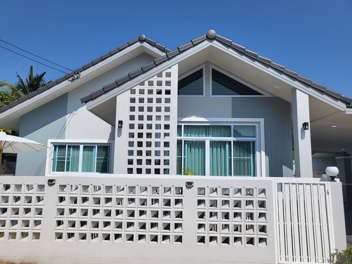 The front exterior of a modern house features a stylish combination of light gray and white walls. Large windows are complemented by a decorative screen and a pitched roof, creating an inviting visual contrast. A welcoming entrance can be seen through the gated fence.