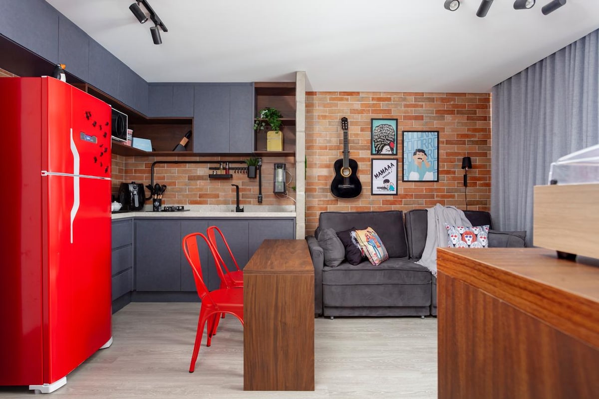 A modern kitchen area features a vibrant red refrigerator and a wooden dining table paired with two red chairs. A cozy gray sofa is positioned nearby, adorned with colorful cushions. Brick accents and artwork add character to the inviting space.