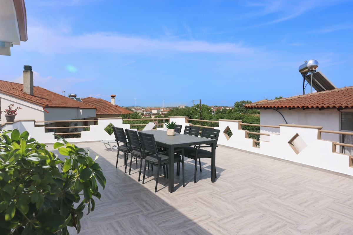 A large private balcony is featured with a spacious dining table surrounded by chairs. Greenery is visible in the foreground, while scenic views of the village and blue sky are seen beyond. The balcony's design includes decorative openings, enhancing the space's openness.