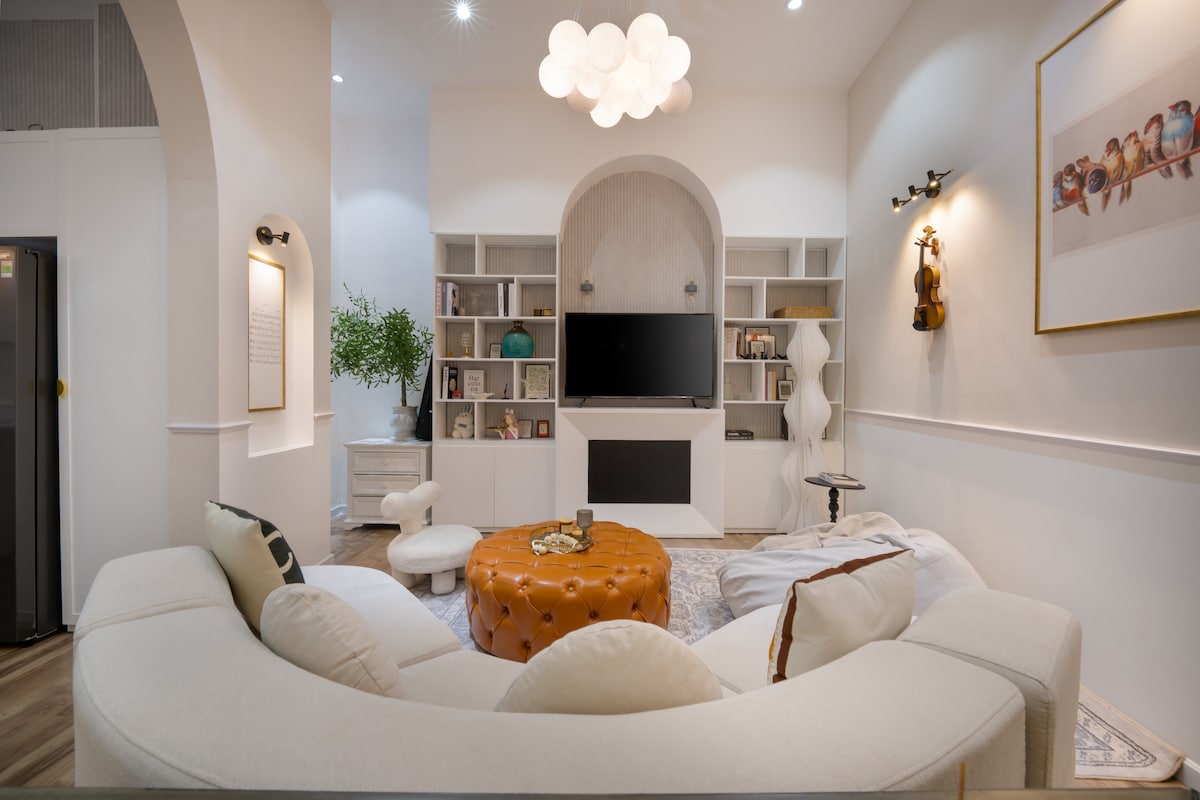 A modern and welcoming living room features a curved sectional sofa and a round leather coffee table. A large television is mounted on a white entertainment unit, surrounded by shelves filled with books and decor. Soft lighting complements the serene atmosphere.