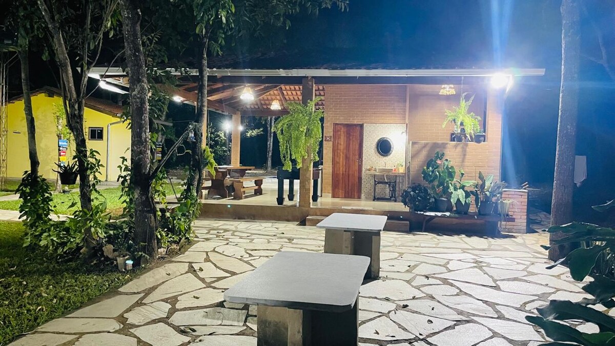 An outdoor patio area is depicted, featuring stone flooring and multiple seating arrangements. A well-lit structure with an open porch is visible in the background, surrounded by greenery and potted plants. A barbecue grill is also present, enhancing the outdoor atmosphere.
