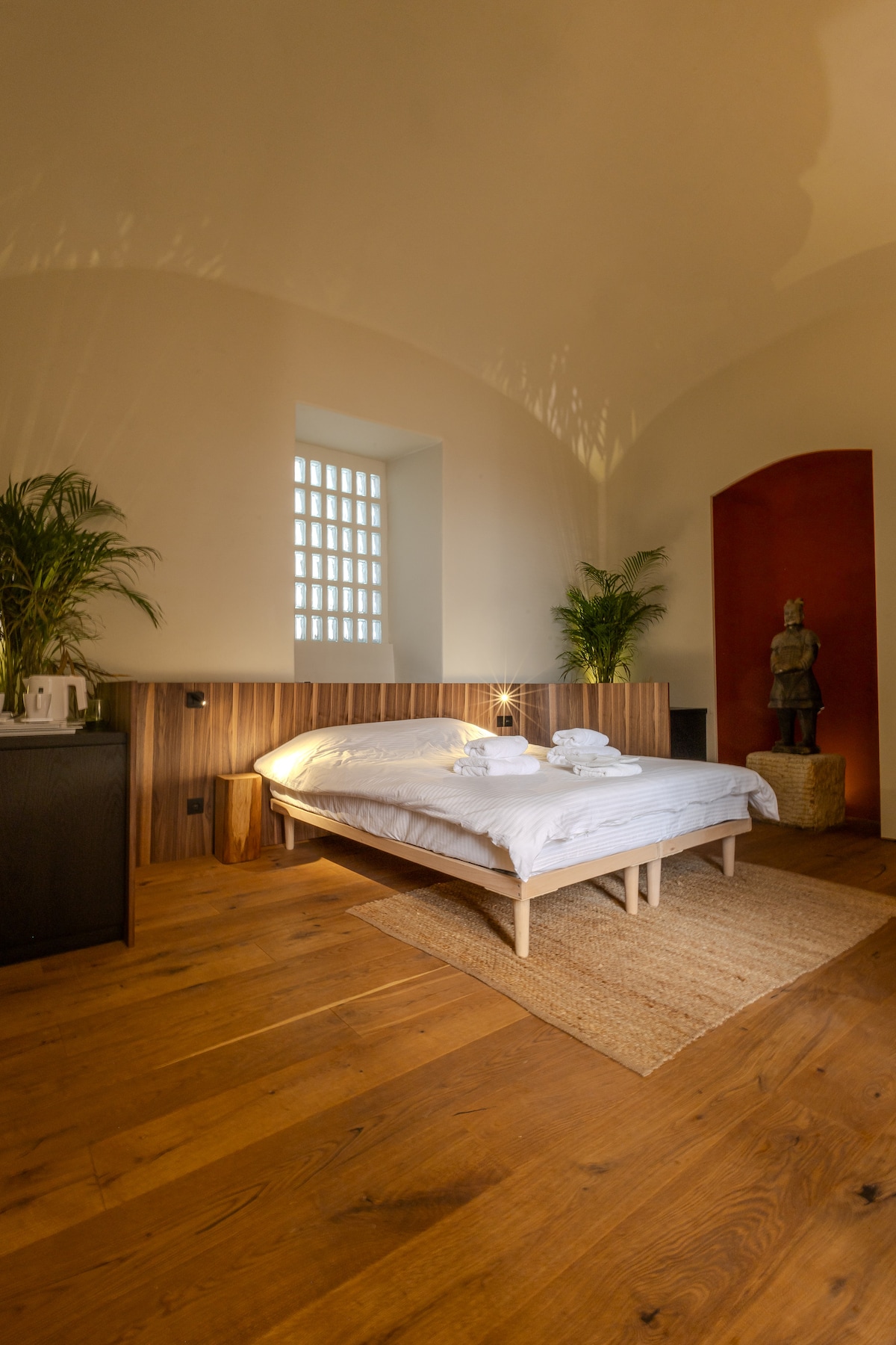 A serene bedroom features a low-profile bed with neatly arranged linens and fluffy towels. Natural light filters through a small window, illuminating wooden flooring and a textured rug. Potted plants add a touch of greenery, while art pieces create a touch of character in the space.