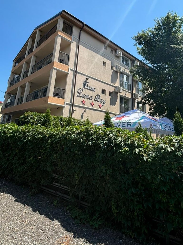 The exterior of Casa Lema Bay is displayed, showcasing a three-story building with a light-colored façade. Balconies adorn the upper levels, while greenery lines the foreground, providing a natural contrast. A colorful awning is visible, indicating an outdoor seating area near the entrance.