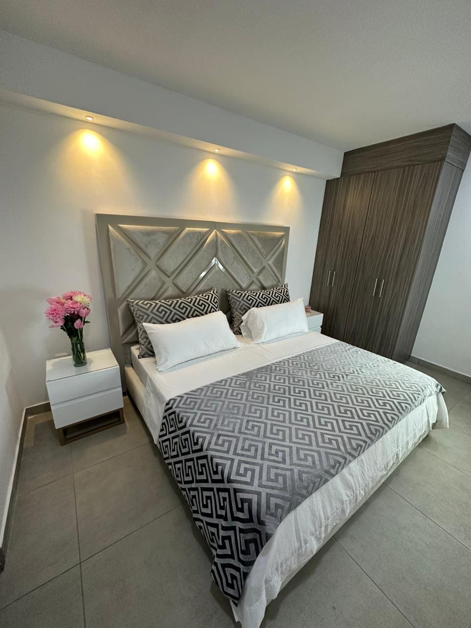 A stylish bedroom is presented with a spacious bed adorned in a patterned grey and white blanket. Soft lighting from the wall fixtures creates a calming ambiance. A bedside table holds a vase of flowers, and a large wardrobe provides ample storage space.