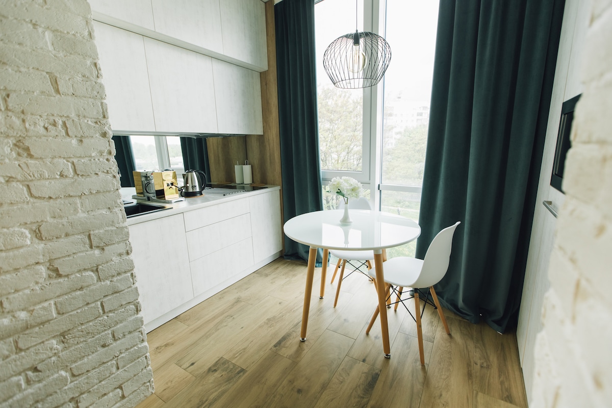 A modern kitchenette is showcased with sleek cabinetry and a minimalist design. A round dining table with two chairs is positioned near a large window, which is adorned with floor-length dark curtains. Natural light filters through, illuminating the warm wooden flooring.