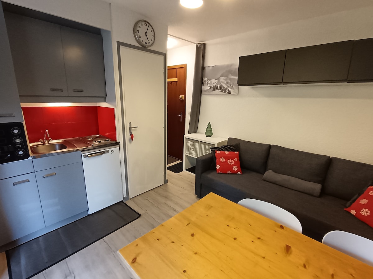 The kitchen area features modern cabinetry with a sink and cooking space, complemented by a vibrant red backsplash. A cozy living space includes a gray sofa adorned with red and white patterned cushions, alongside a wooden dining table and minimalist white chairs.