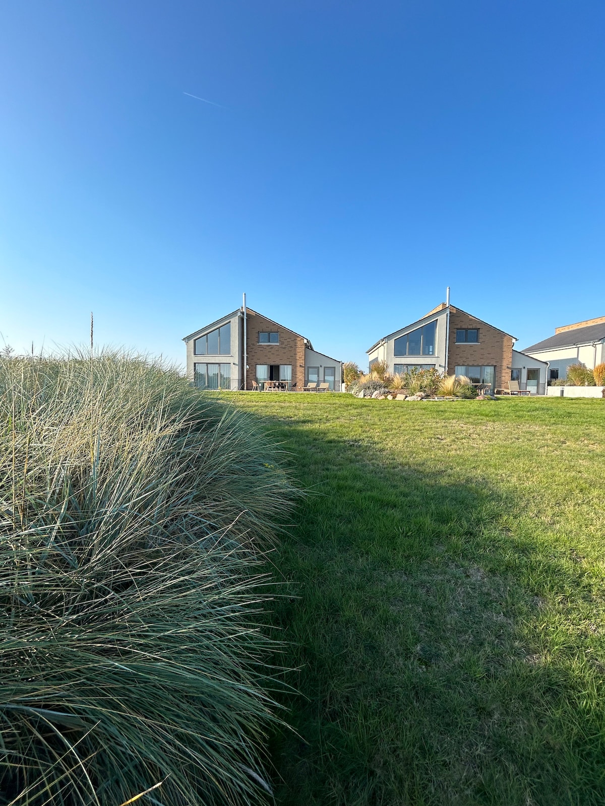 Two charming vacation homes are set against a clear blue sky. Grass surrounds the properties, and lush greenery adds to the natural setting. Each house features large windows, reflecting a modern architectural style, while small outdoor seating areas can be seen in the distance.
