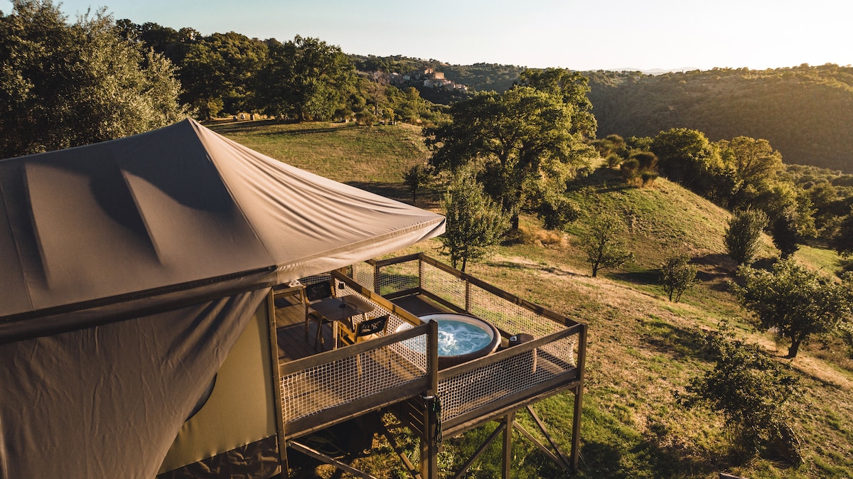 The tent structure is positioned on a wooden deck, featuring a small hot tub surrounded by a scenic landscape. Rolling hills and trees create a natural backdrop, with the setting sun casting a warm golden light across the area.