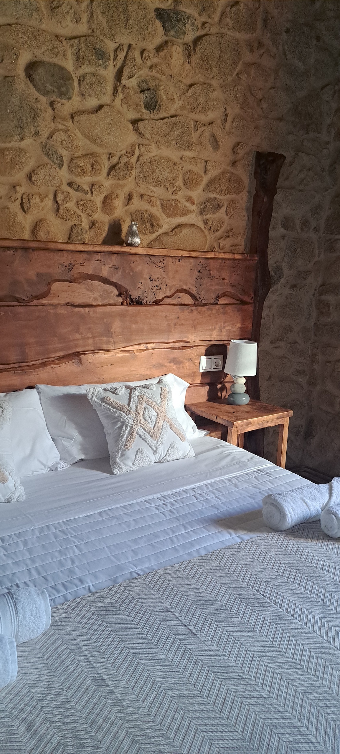 A cozy bedroom is highlighted by a rustic stone wall and a wooden bed frame. A soft, light blanket rests on the spacious bed, accompanied by neatly rolled towels. A bedside table with a lamp provides gentle illumination, enhancing the tranquil setting.