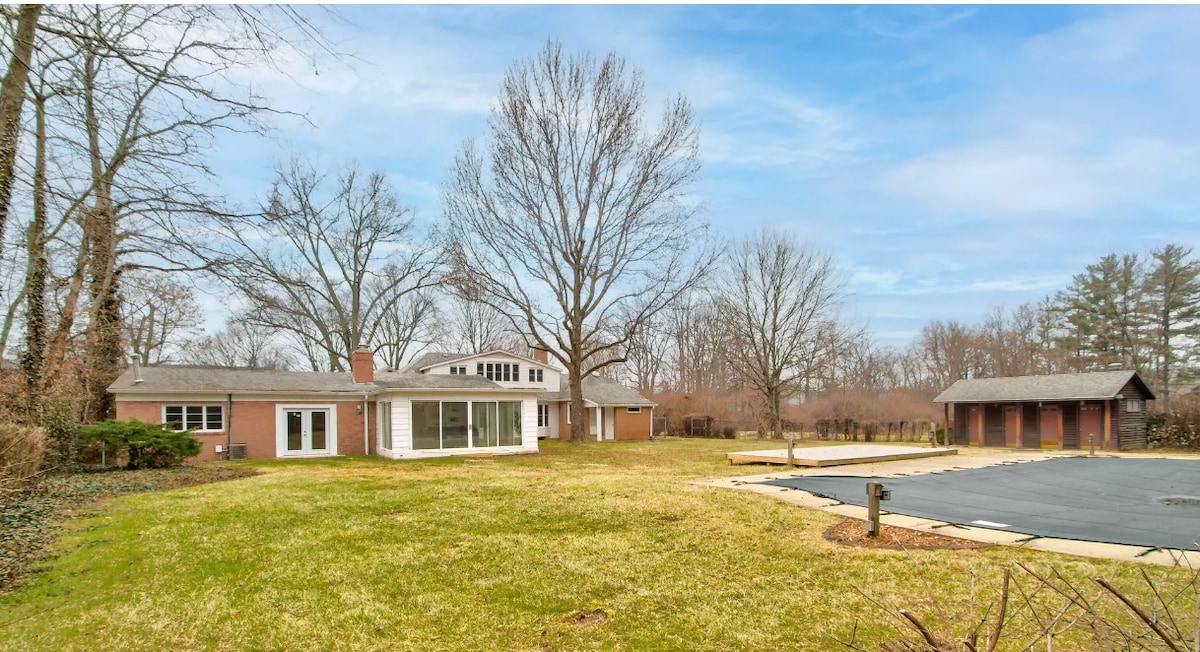 A spacious outdoor area is depicted, featuring a large lawn surrounded by trees. The house showcases a combination of brick and large glass panels, providing a view of expansive green spaces. A paved area is visible to the right, adjacent to a utilitarian structure.