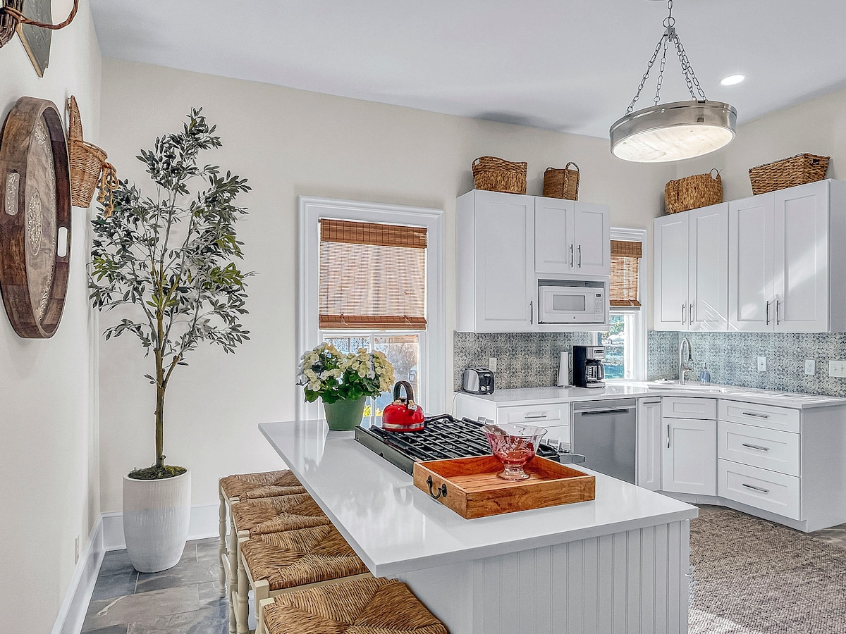 The kitchen features modern white cabinetry and a spacious island with seating for four. A professional gas stove and stainless steel appliances are visible, complemented by natural light from the windows adorned with bamboo shades. Decorative baskets and a potted plant add a touch of warmth.