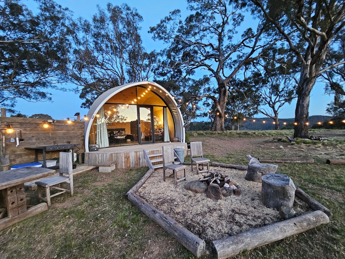 A cozy outdoor area features a firepit surrounded by wooden seating. The tiny home, with its curved design, showcases large glass doors that open to the inviting space. String lights overhead enhance the connection to the natural surroundings, including the nearby trees.