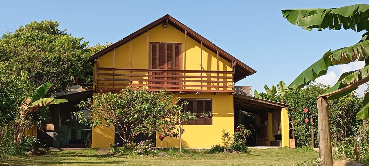 The exterior of a yellow chalet is prominently displayed, featuring a wooden balcony and large windows. Lush greenery surrounds the structure, with banana plants and flowering bushes enhancing the landscape. A covered porch area is visible, providing shaded outdoor space.