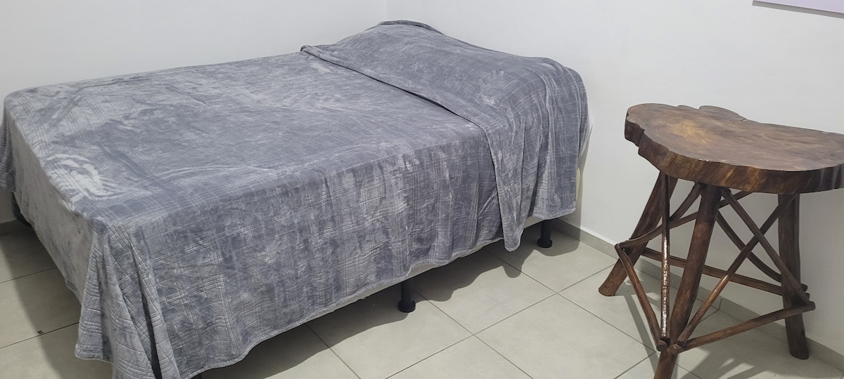 A spacious bed draped with a soft gray coverlet is positioned beside a uniquely shaped wooden side table. The room features tiled flooring and light-colored walls, creating a simple, clean environment.