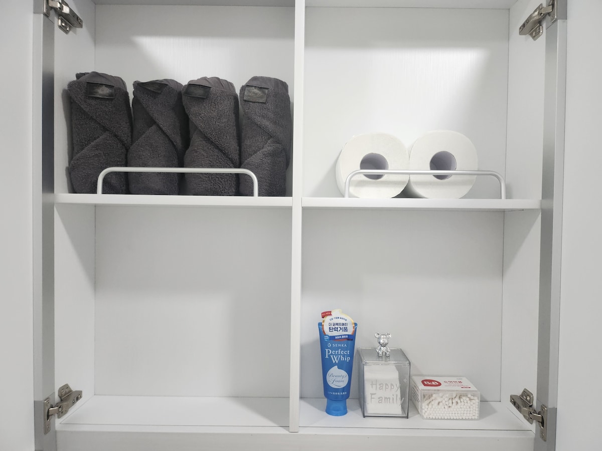 A neatly organized cupboard displays three rolled towels in dark fabric, placed on a shelf. Above them are two rolls of toilet paper. A bottle of mouthwash and a small container labeled 'Happy Family' are positioned on the bottom shelf, enhancing the functional layout.