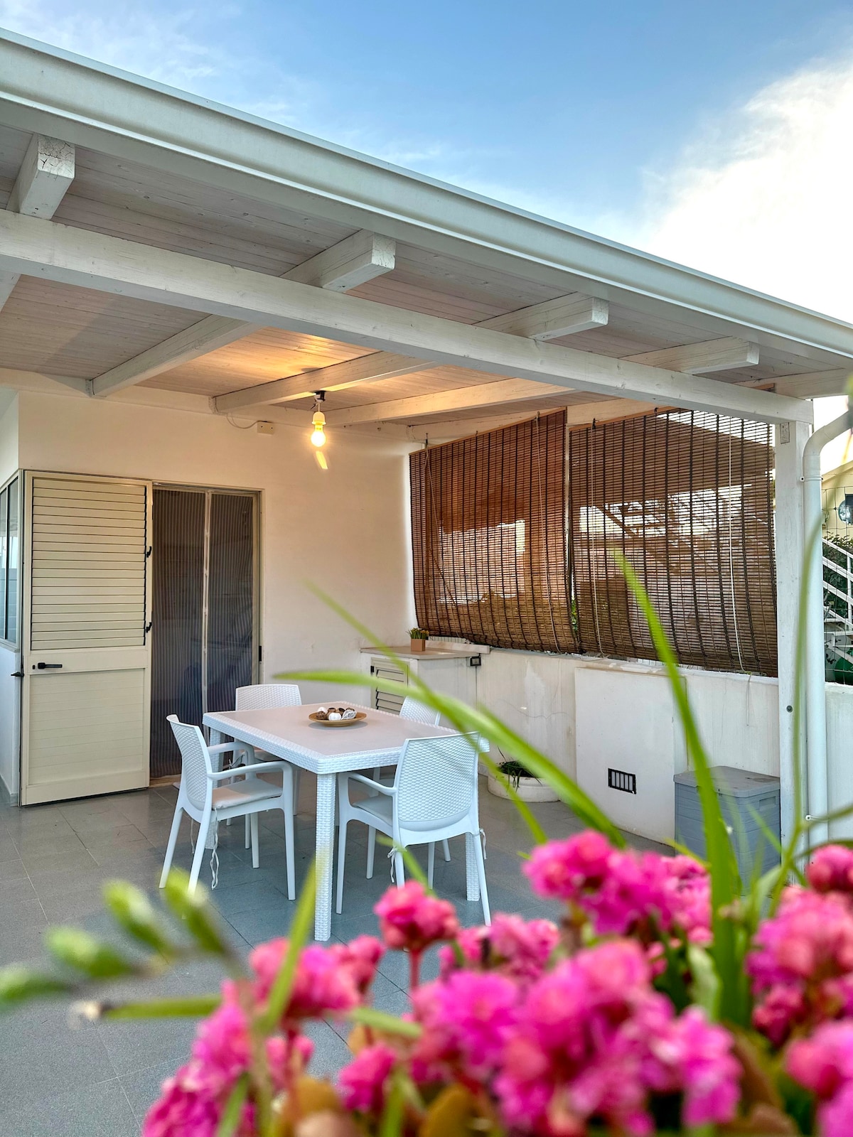 An outdoor veranda is featured, showcasing a dining table surrounded by white chairs. Natural light filters through bamboo shades, while colorful flowers are positioned in the foreground. The space is designed for relaxation and dining, with a ceiling fan for comfort.
