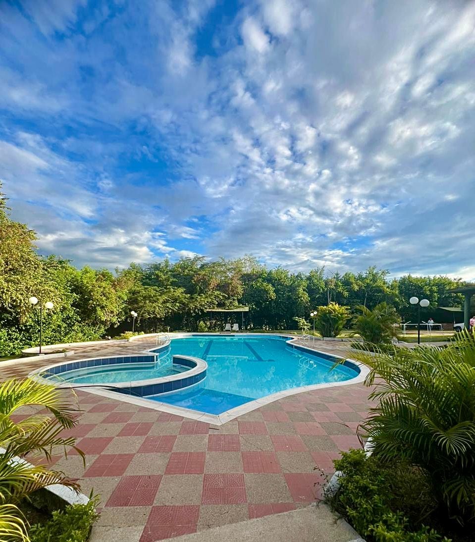 A spacious outdoor pool area is surrounded by lush greenery, featuring a large swimming pool with a shallow section and a jacuzzi. The patio is paved with red and gray tiles, and several light fixtures are positioned around the space, creating a welcoming atmosphere.